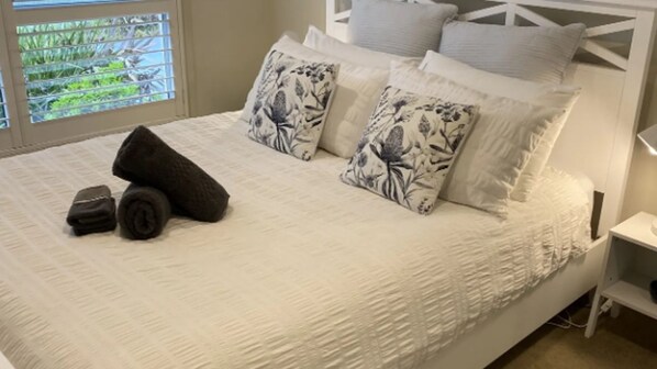 A neatly made bed with white bedding and floral-patterned pillows, a small bedside table, and a window with a view of greenery.