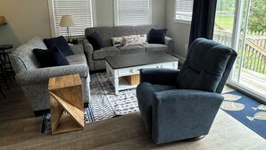 Ett vardagsrum med en grå soffa, en blå fåtölj, ett träbord och en takfläkt.