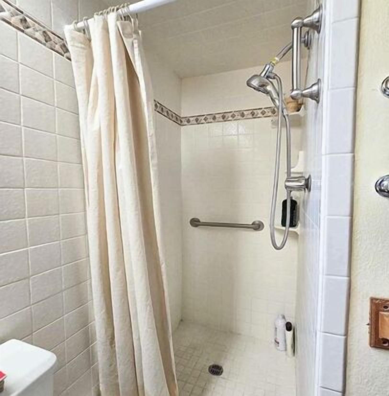 A small, tiled shower room with a showerhead, a handrail, and a curtain.