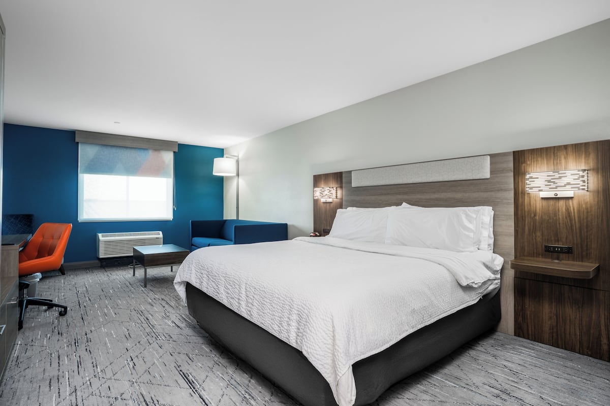 A modern hotel room with a large bed, a desk, a chair, and a window with blinds.