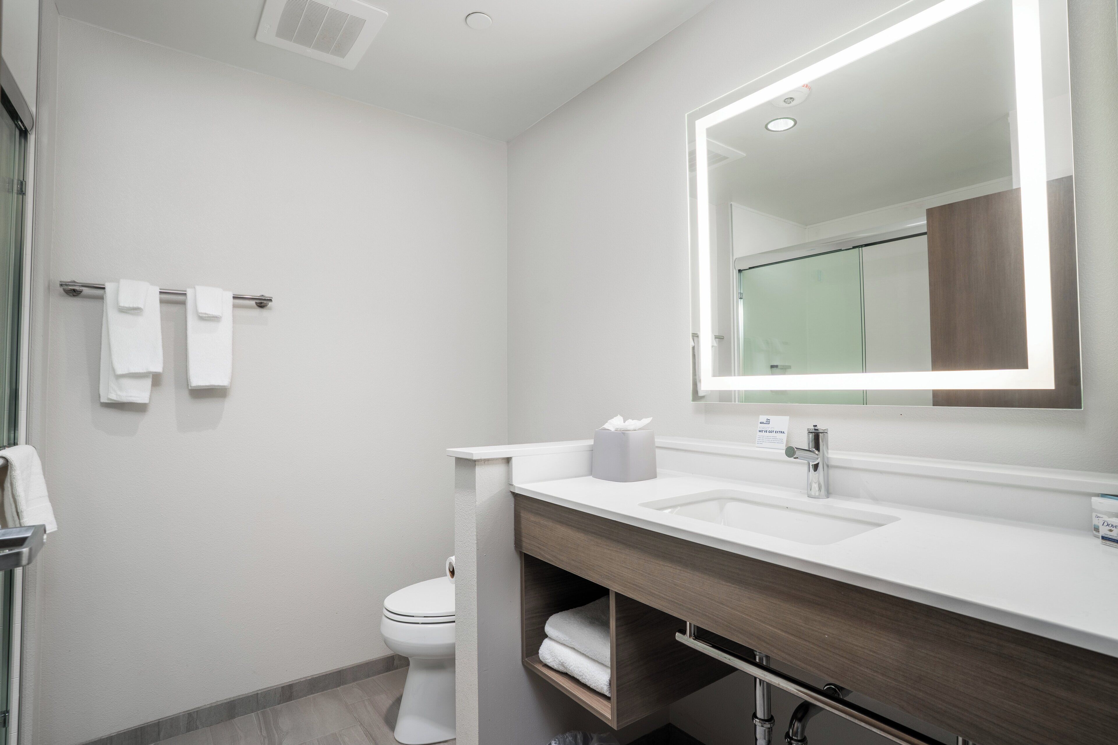 Et moderne badeværelse med hvid vask, spejl og toilet. Der er håndklæder på en holder og en tissueske på bordet.