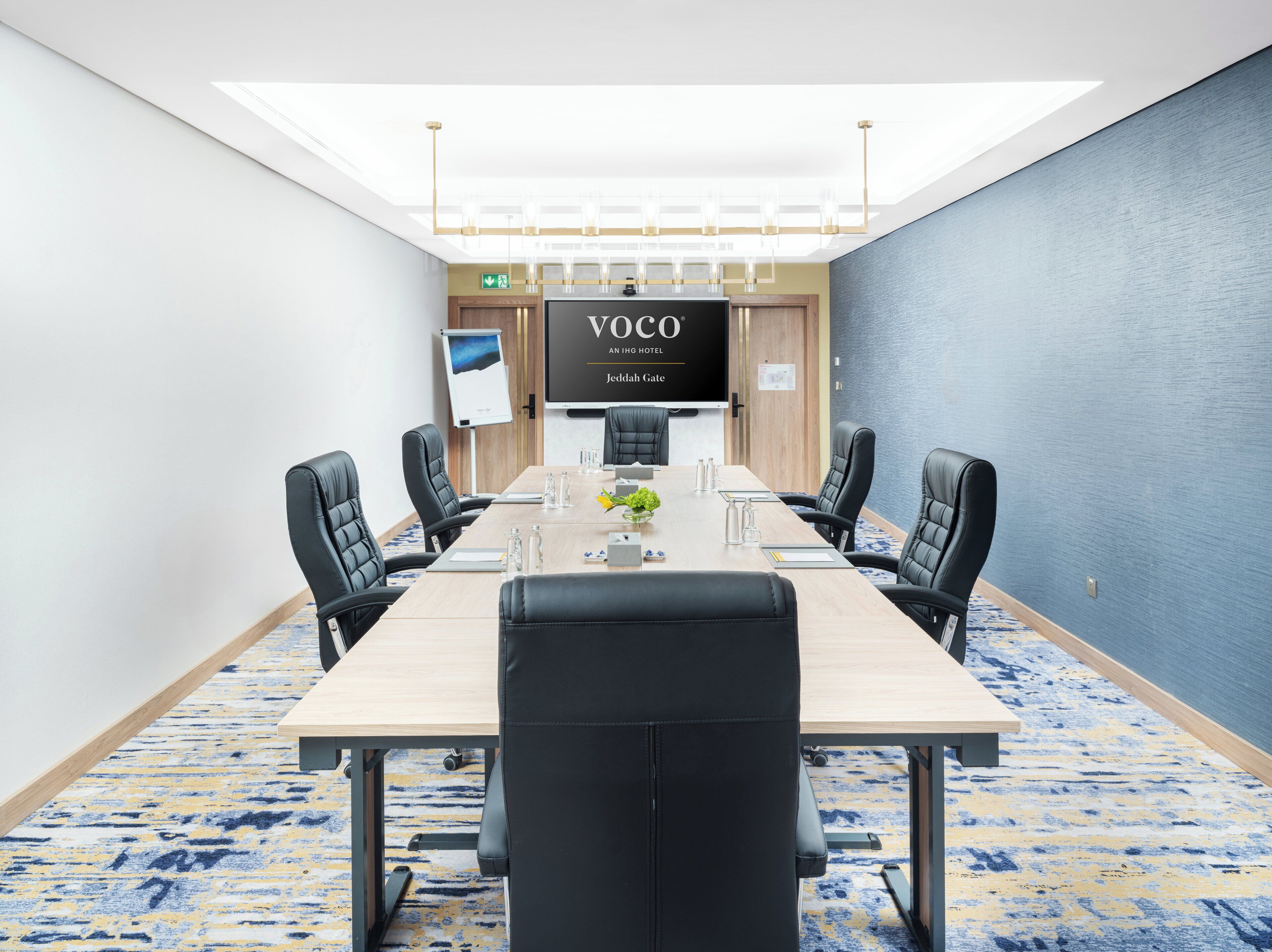 A conference room with a long table, black leather chairs, and a large screen displaying "VOCO AN HOTEL Jeddah Gate".