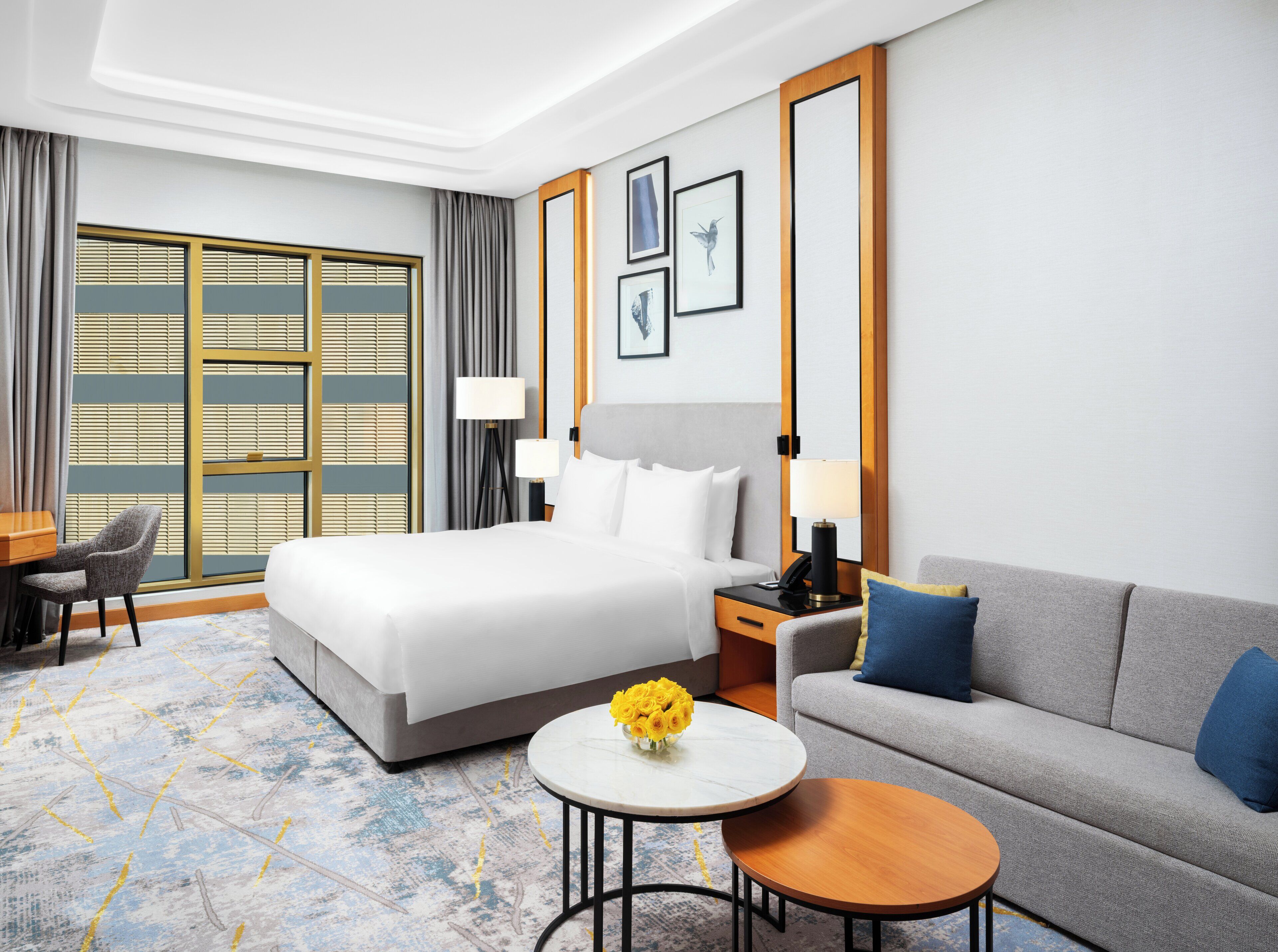 Une chambre d’hôtel avec un lit, un canapé, une table basse ronde, un bureau et une chaise. Il y a des rideaux, une lampe et des tableaux encadrés au mur.