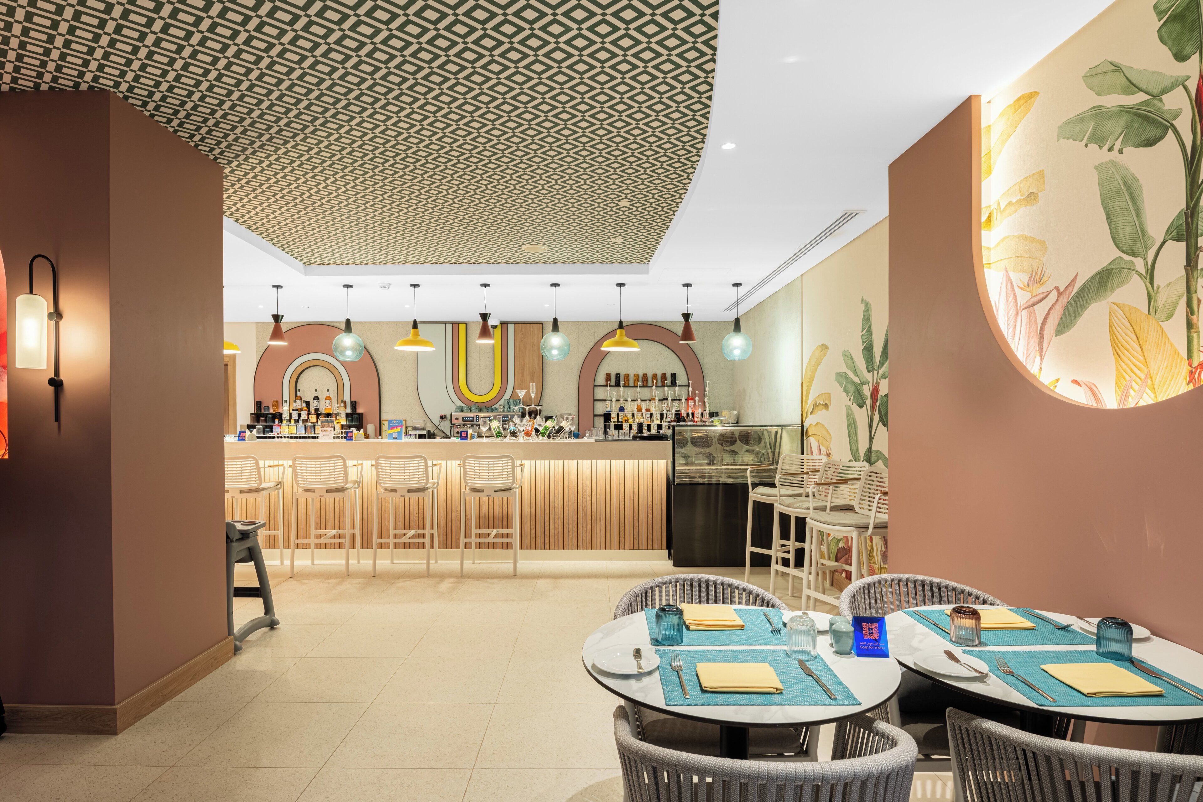 A modern restaurant with a bar area, round tables with chairs, and a patterned ceiling.