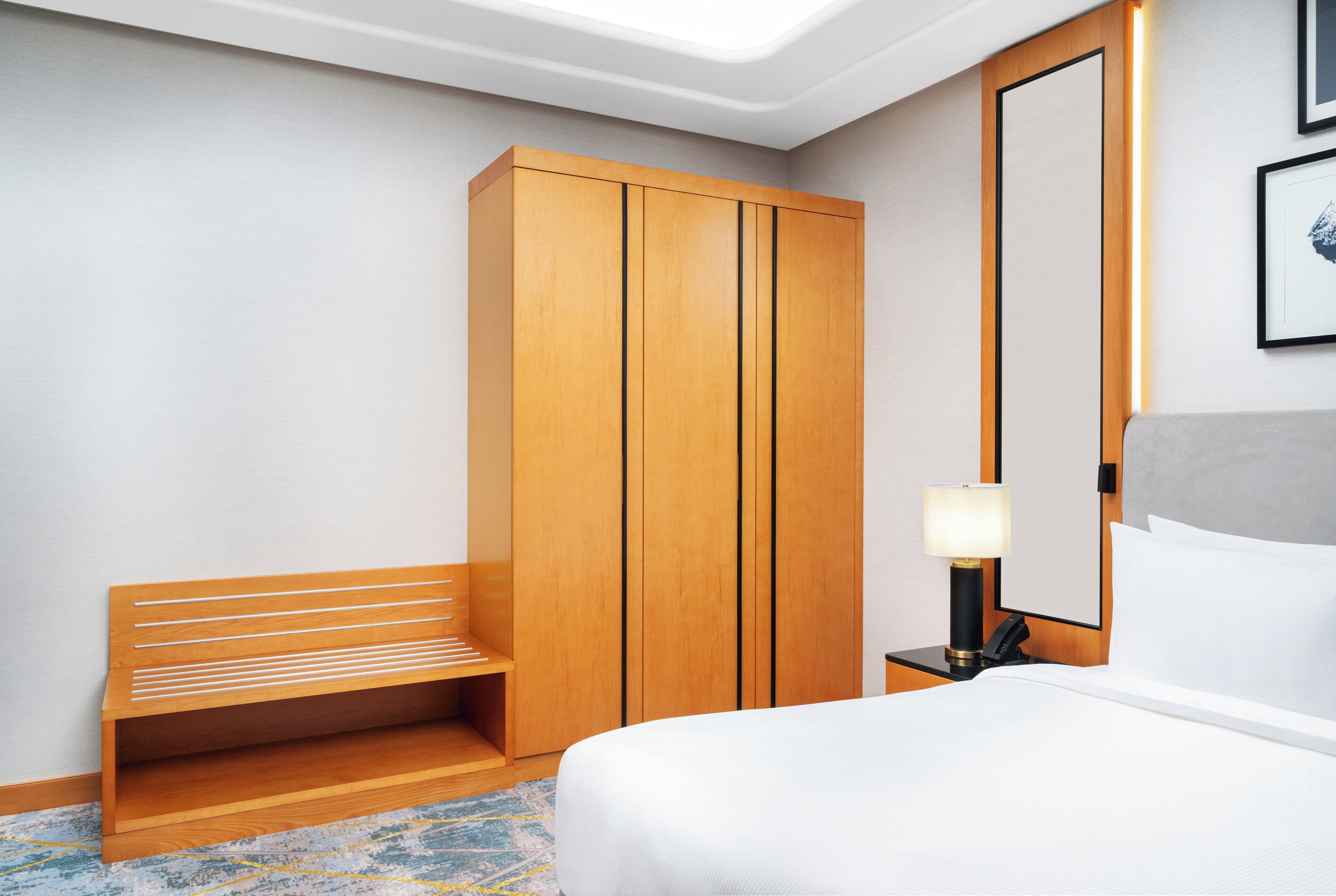 A hotel room with a wooden wardrobe, a bench, a bedside table with a lamp, and a bed with white bedding.