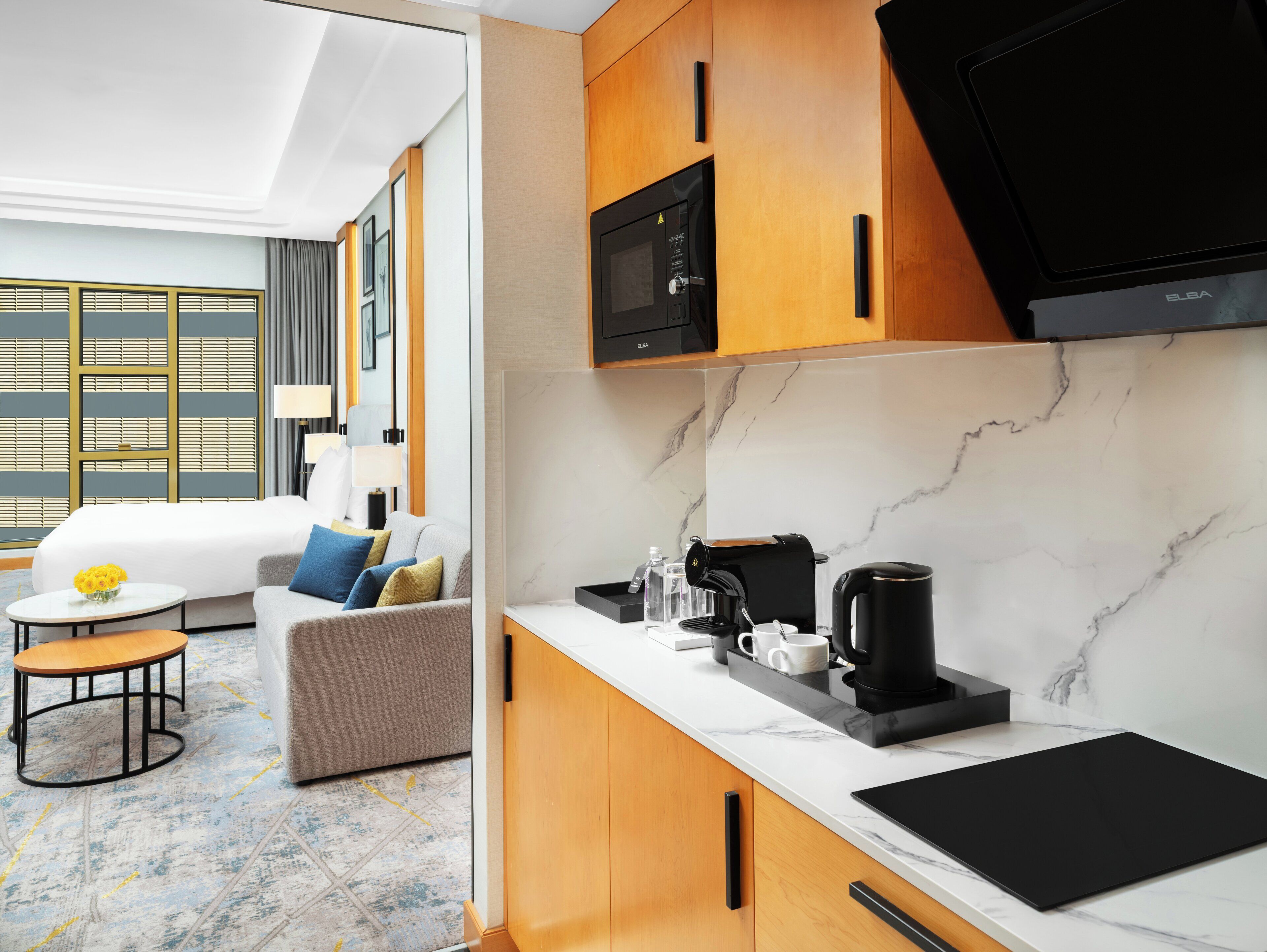 A modern hotel room with a sofa, a small round table with a vase of flowers, a coffee maker, and a TV.