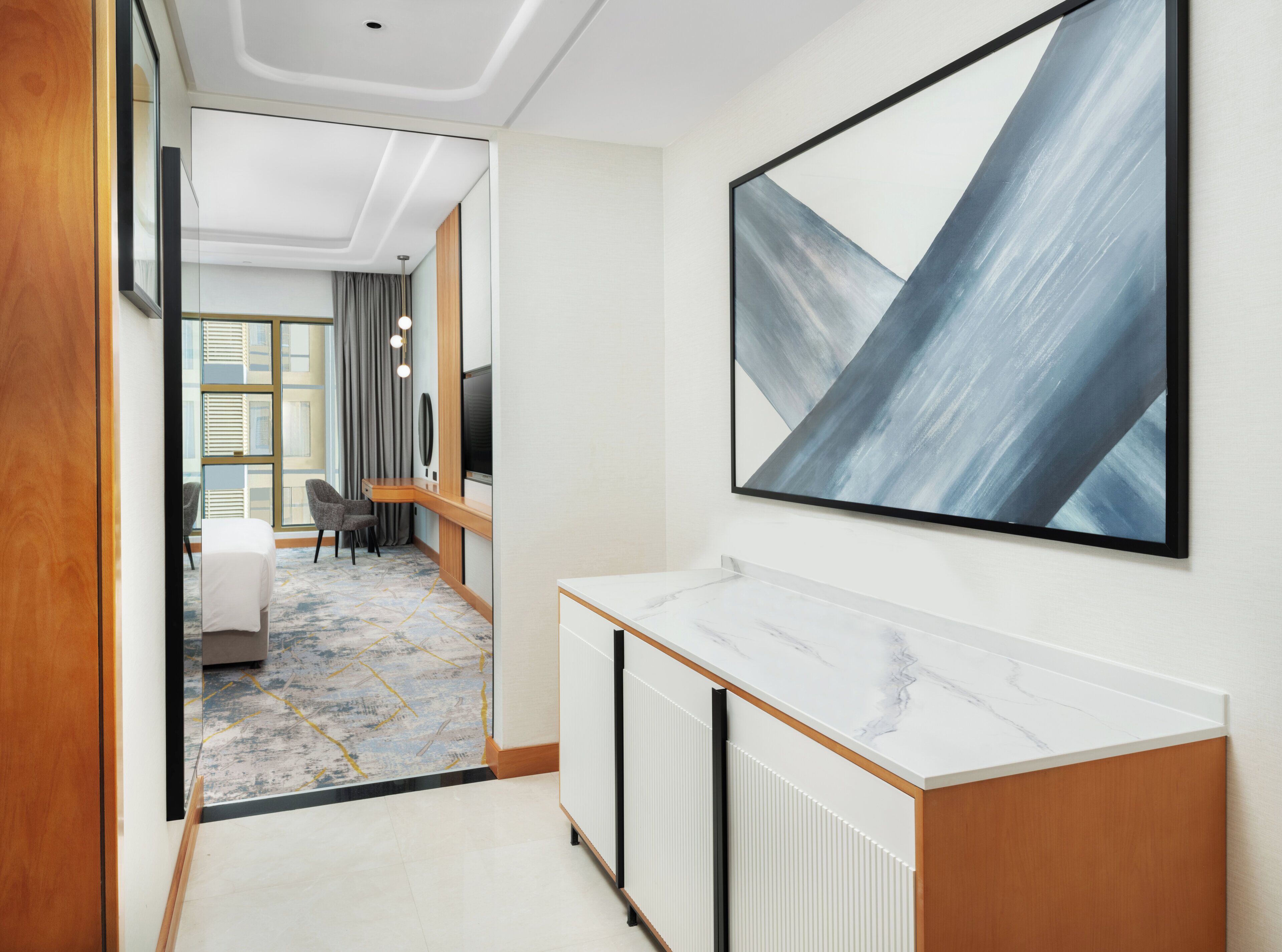 A modern hotel hallway with a marble reception desk, a large abstract painting, and a view into a room with a bed and a desk.