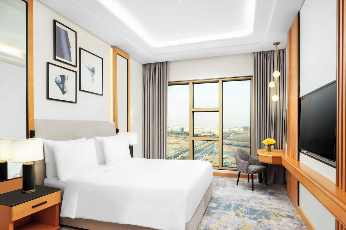 A hotel room with a large bed, a desk with a chair, a TV, and a city view through the window.