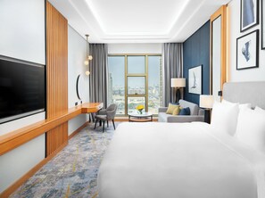 A hotel room with a large bed, a TV, a desk, a chair, a sofa, a lamp, and a view of the city. - voco Jeddah Gate by IHG (Jeddah)