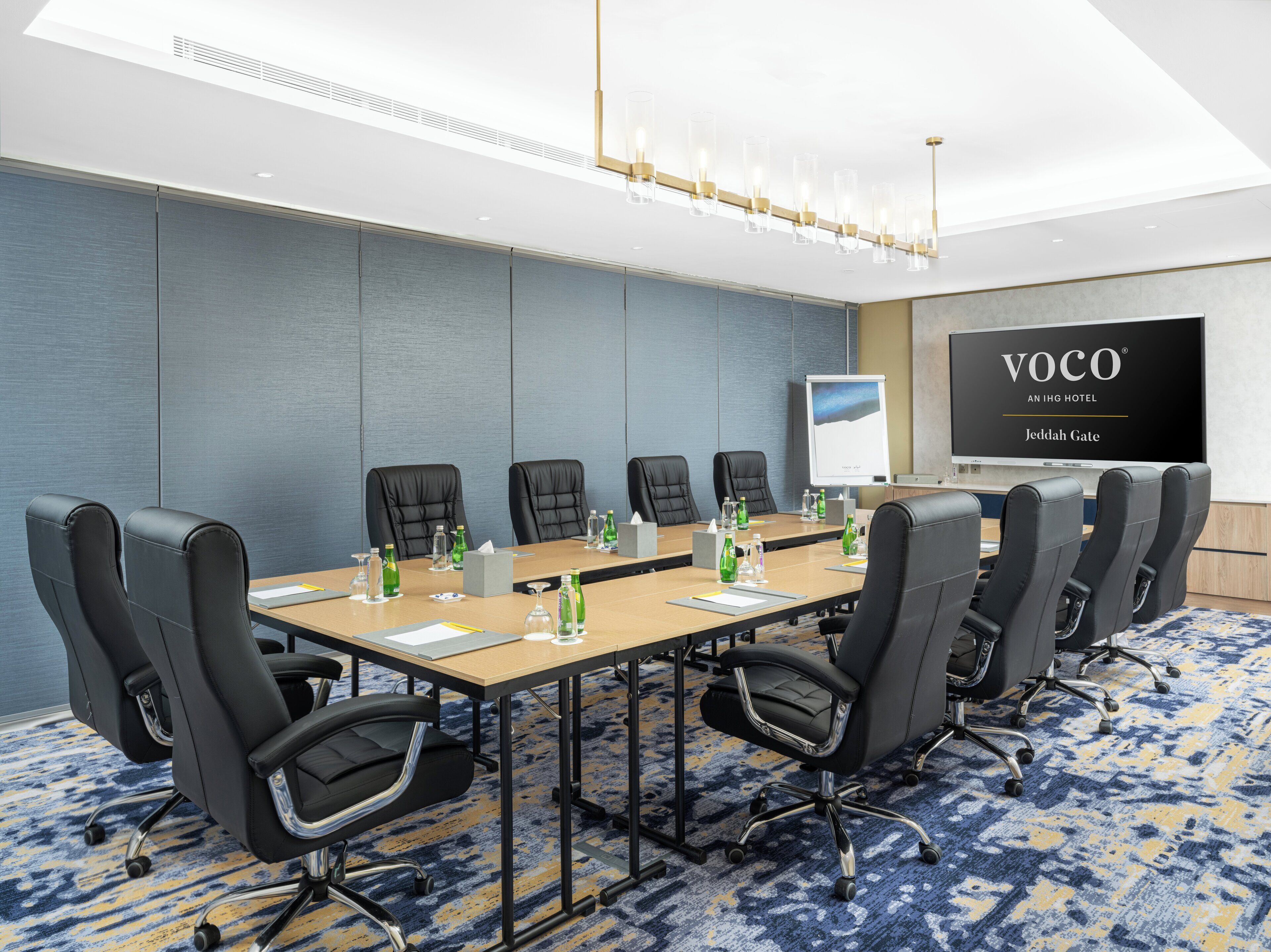 Uma sala de conferências com uma mesa comprida, cadeiras de couro preto, um quadro branco e uma tela grande exibindo "VOCO AN IHG HOTEL Jeddah Gate".
