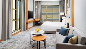 A hotel room with a bed, a sofa, a desk with a TV, a small table with flowers, and large windows with curtains. - voco Jeddah Gate by IHG (Jeddah)