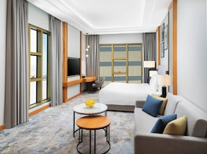 A hotel room with a bed, a sofa, a desk with a TV, a small table with flowers, and large windows with curtains. - voco Jeddah Gate by IHG (Jeddah)