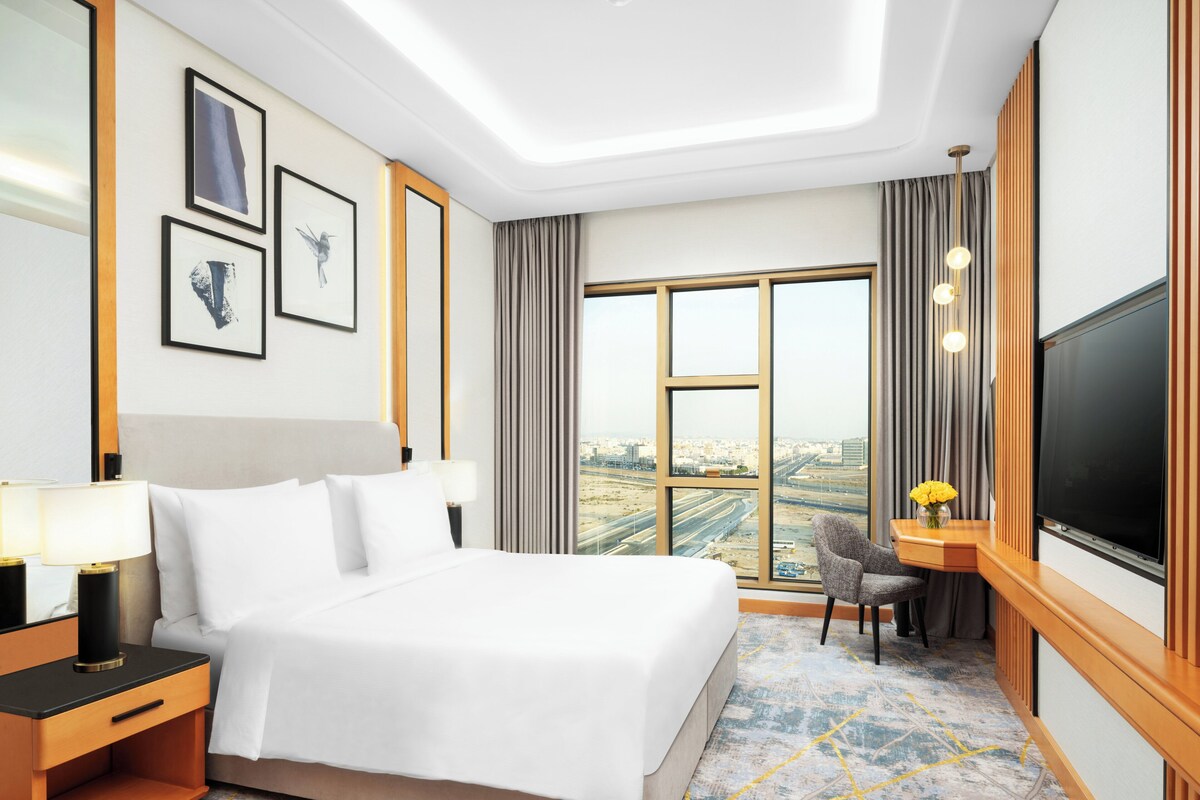 A hotel room with a large bed, a desk with a chair, a TV, and a city view through the window.