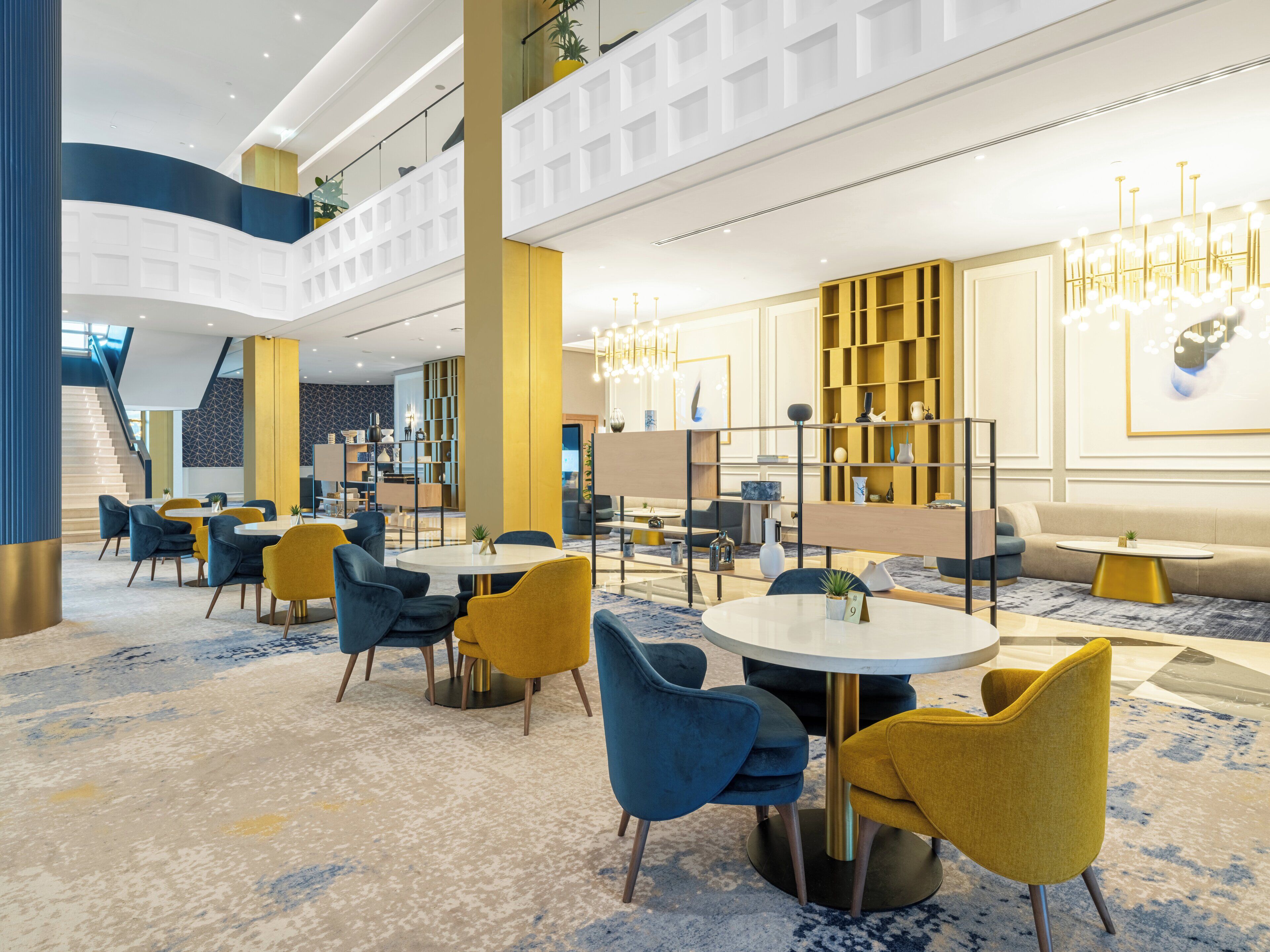 Een moderne hotellobby met ronde tafels, blauwe en gele stoelen en een trap.