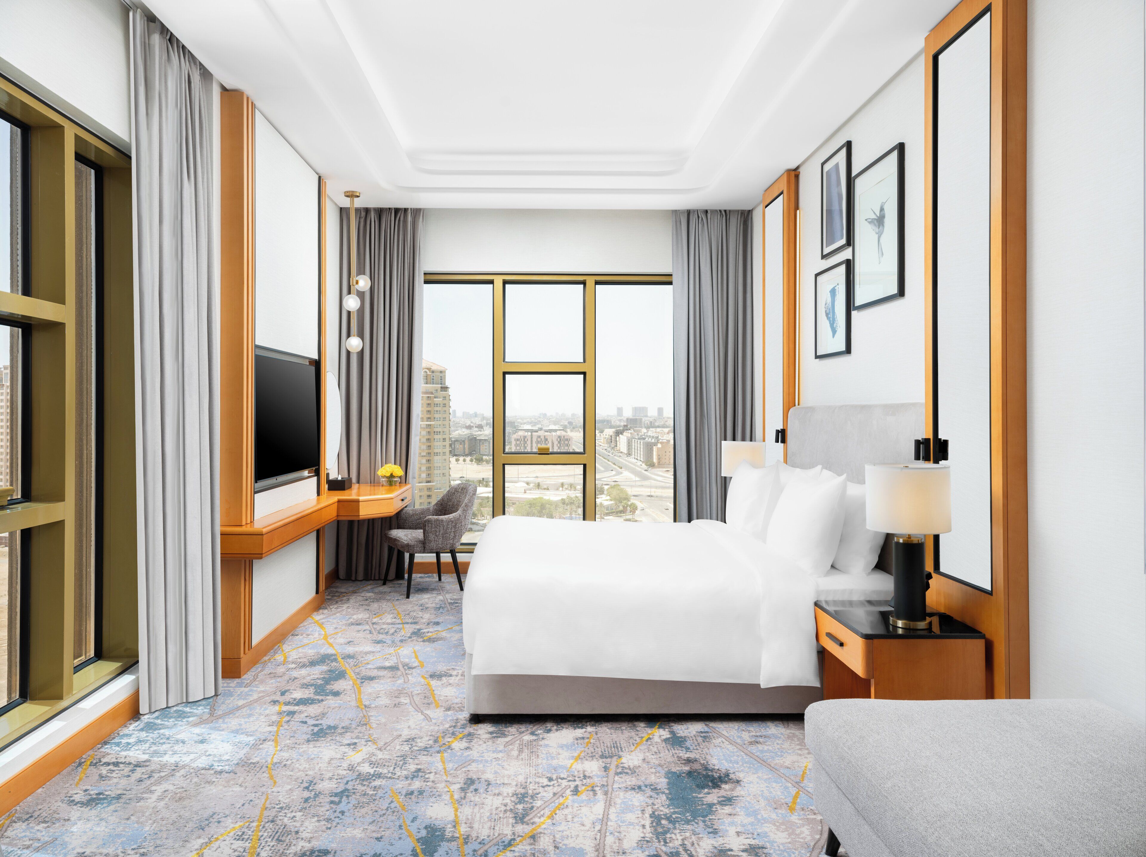 Habitación de hotel con una cama grande, un escritorio con televisor, una silla, una lámpara y vistas a la ciudad.