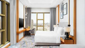 Habitación de hotel con una cama grande, un escritorio con televisor, una silla, una lámpara y vistas a la ciudad.