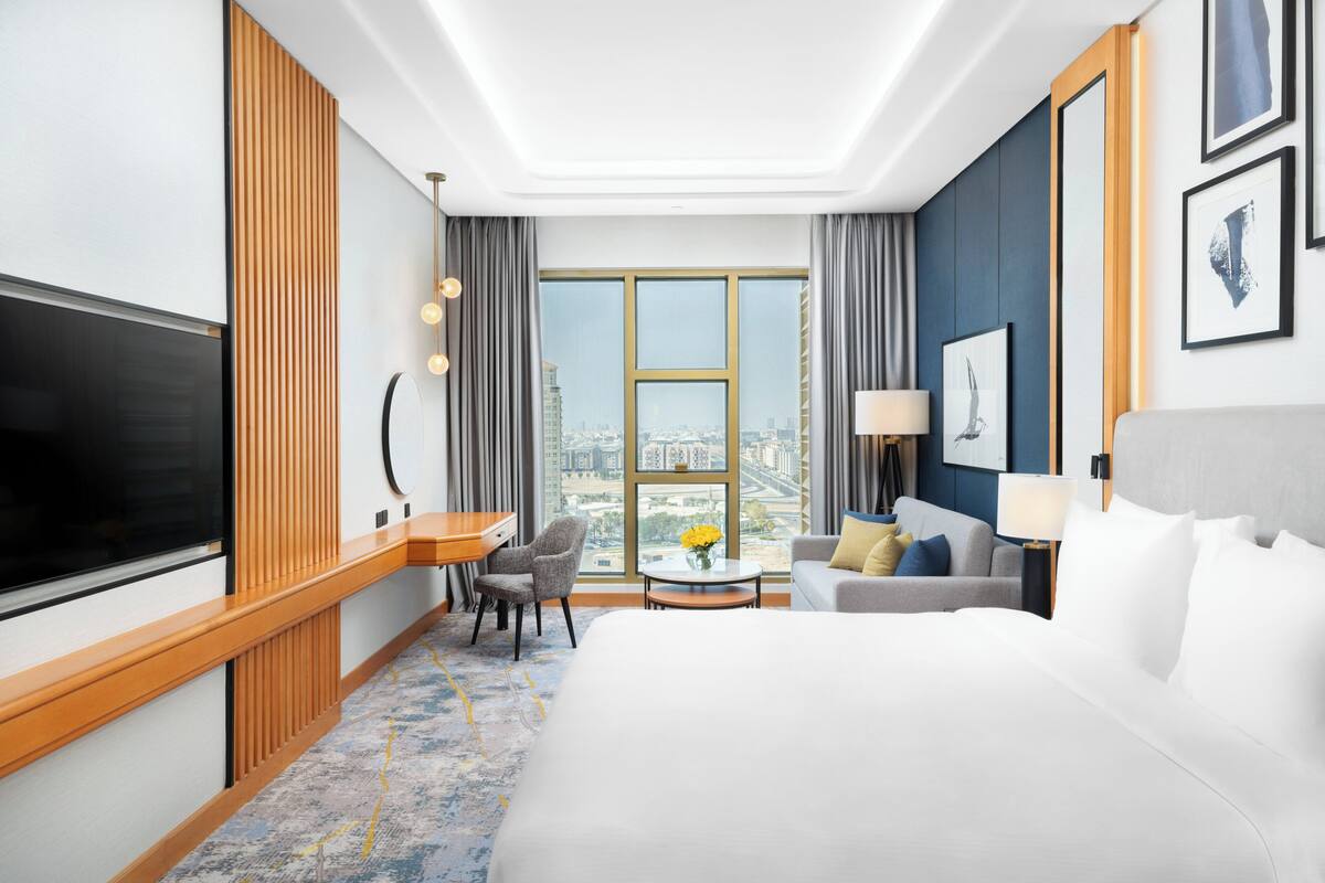 A hotel room with a large bed, a TV, a desk, a chair, a sofa, a lamp, and a view of the city.