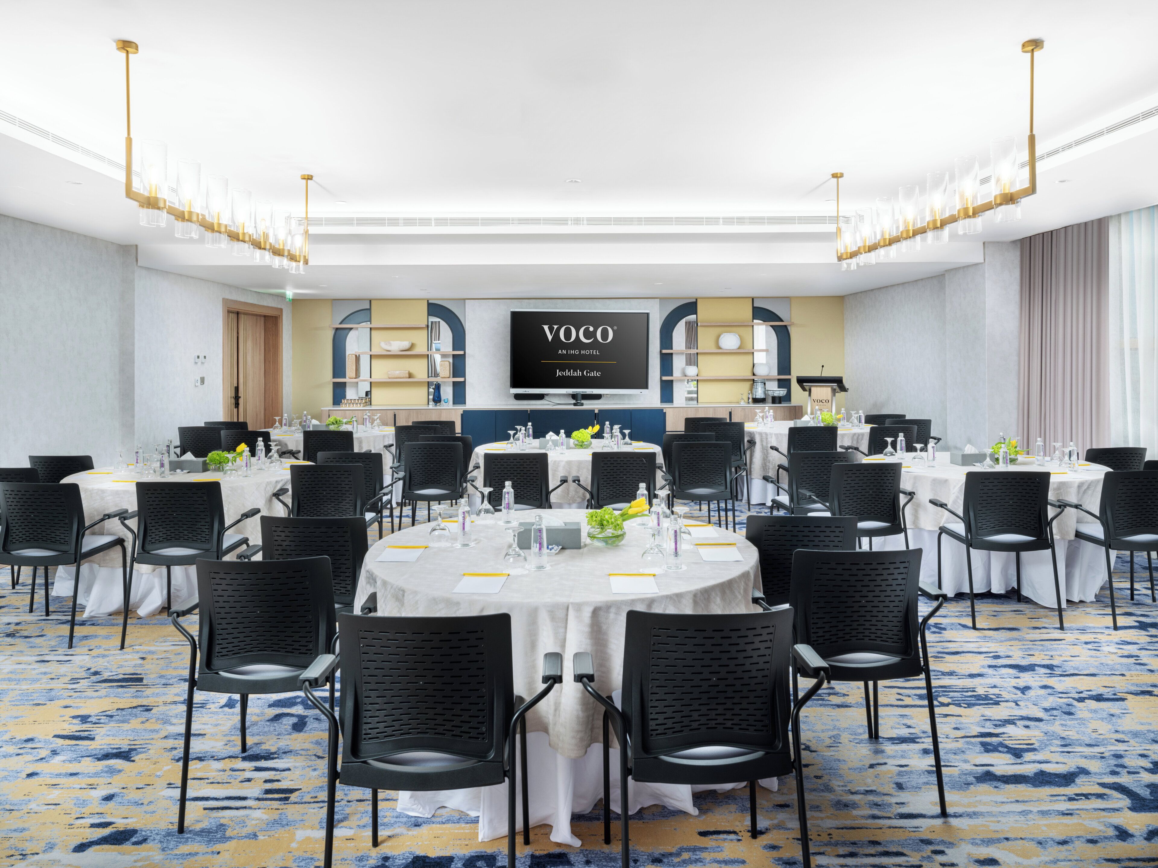 Uma sala de conferências com mesas redondas, cadeiras pretas e uma tela grande exibindo "YOCO AN IHG HOTEL Jedediah Gate".