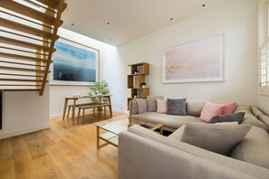 Uma sala de estar moderna com escada de madeira, mesa de centro de vidro, sofá bege e mesa lateral de madeira.