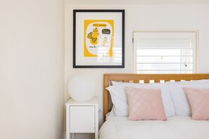 Cama bem arrumada com almofadas brancas e rosa, um quadro na parede e uma mesa de cabeceira com abajur branco.