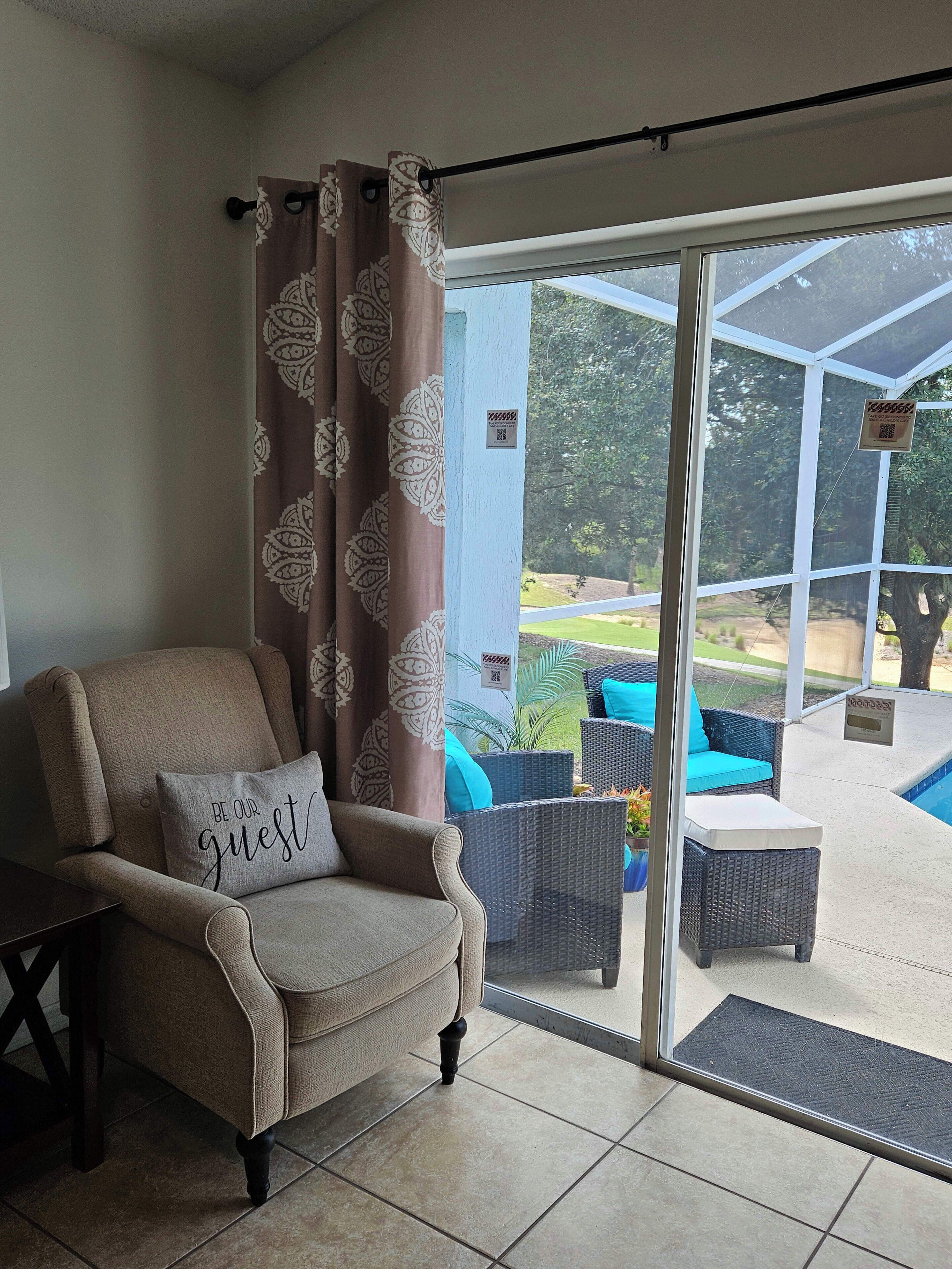 A room with a beige armchair, a cushion with "BE OUR guest" text, a glass door leading to an outdoor area with wicker furniture and a pool.