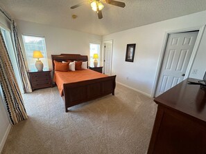 Une chambre à coucher comprenant un lit, une table de chevet, une commode, un ventilateur de plafond et une porte.