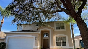 A two-story house with a beige exterior, a white garage door, and a front entrance with a small porch. - Plenty of Sunshine, Southern Dunes Pool Home (Haines City)