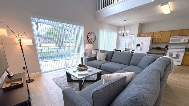 A modern living room with a grey sectional sofa, a coffee table, and a floor lamp. There is a dining area with a table and chairs, and a kitchen with wooden cabinets and a refrigerator. - Plenty of Sunshine, Southern Dunes Pool Home (Haines City)