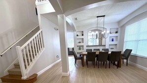 A spacious dining area with a high ceiling and modern chandelier, a staircase with white railings, and a view of the kitchen area. - Plenty of Sunshine, Southern Dunes Pool Home (Haines City)