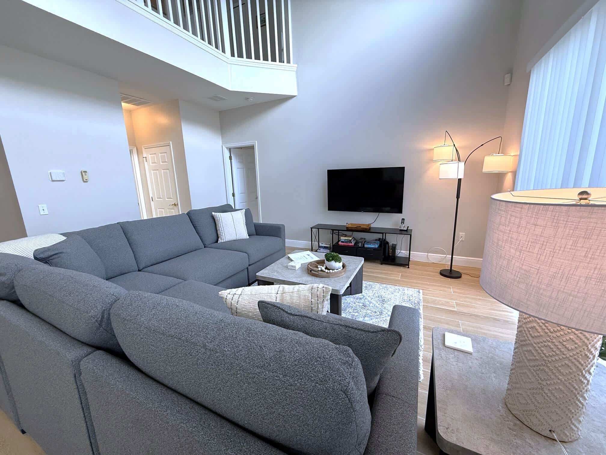 Un salon moderne avec un canapé d’angle gris, une table basse, un téléviseur à écran plat et une lampe.