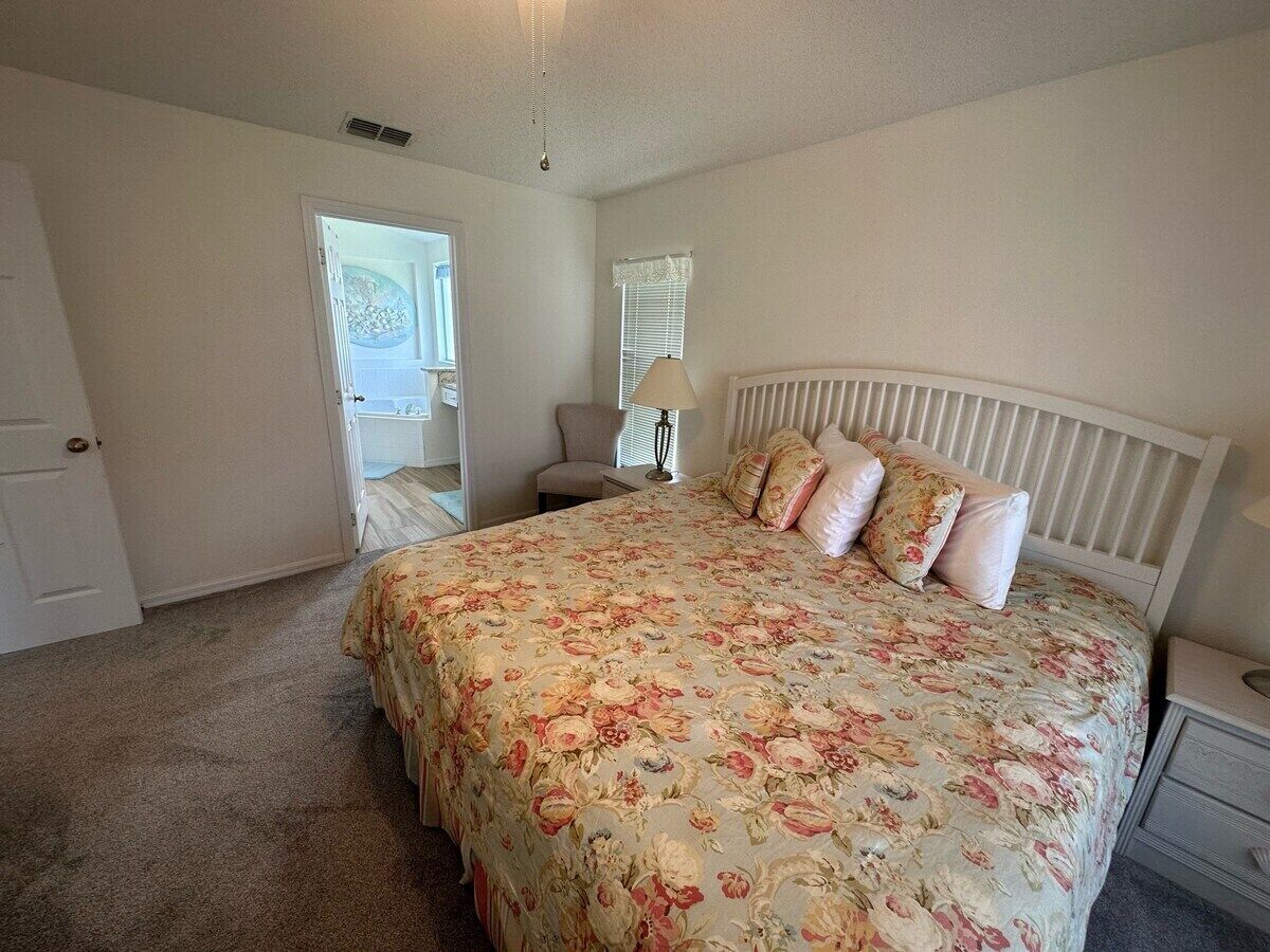 A bedroom with a large bed, a nightstand, a chair, and a door leading to another room.