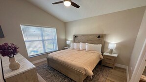 Une chambre à coucher comprenant un lit, des tables de chevet, une commode, un ventilateur de plafond, une fenêtre avec des stores et un plancher en bois.