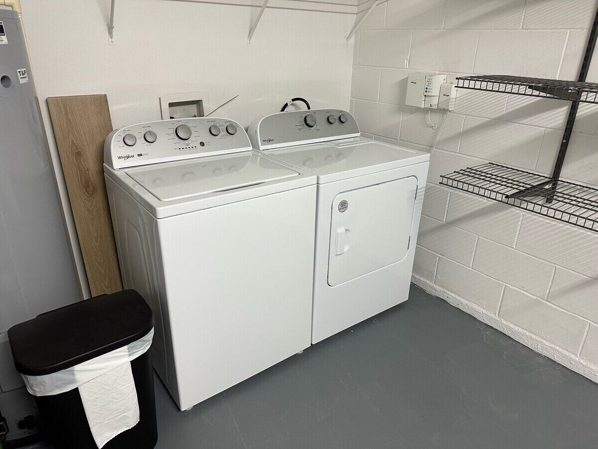 Une buanderie équipée d’un lave-linge et d’un sèche-linge à chargement frontal, d’une étagère en métal et d’une poubelle.