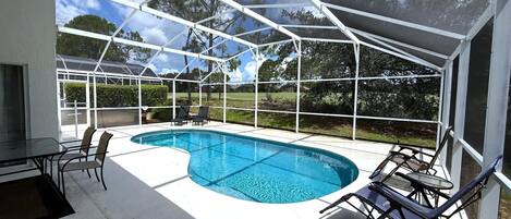 A covered outdoor swimming pool with a glass enclosure, surrounded by chairs and a table.