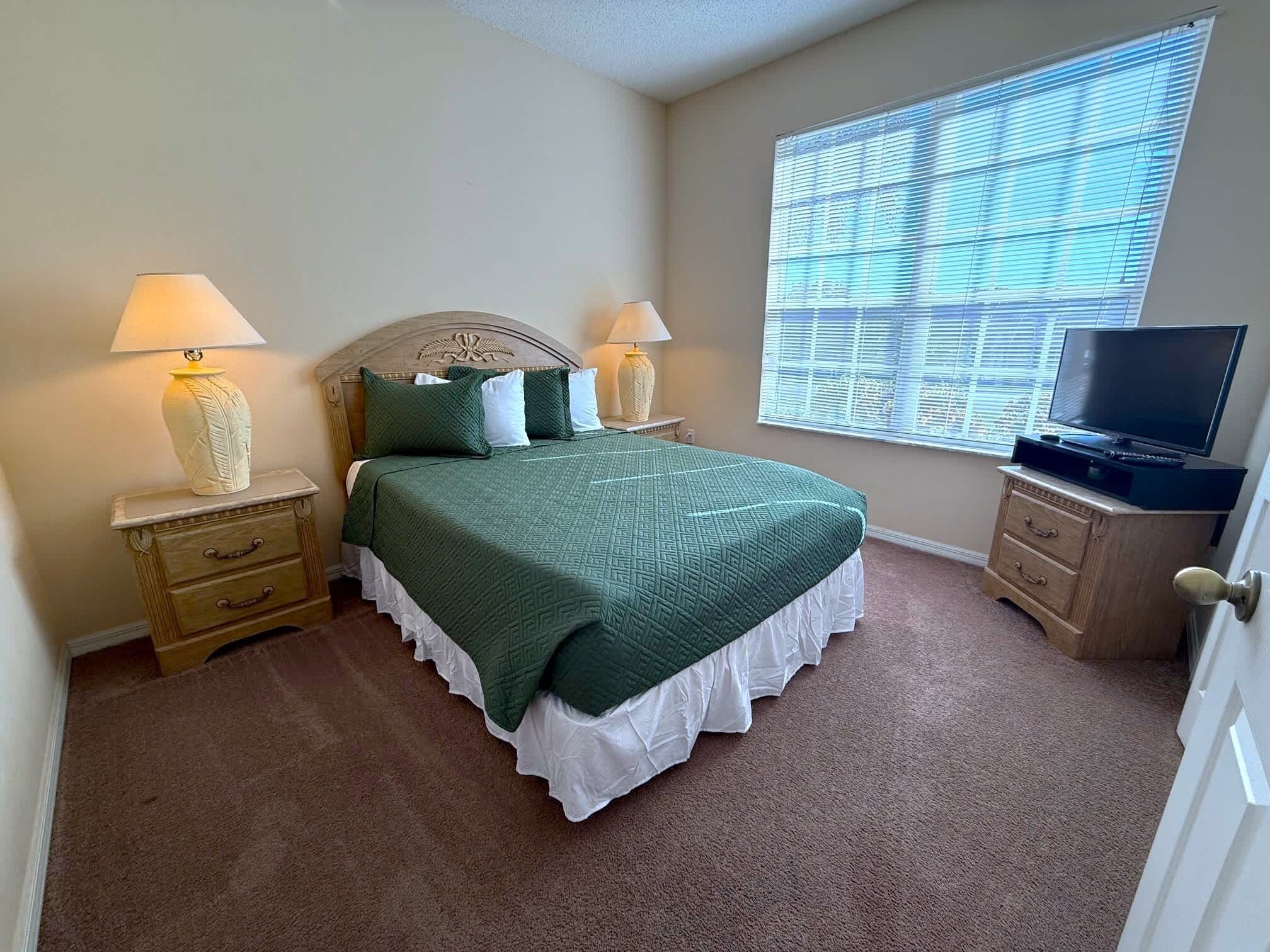 A bedroom with a bed, two bedside tables with lamps, a dresser, a television, and a large window with blinds.
