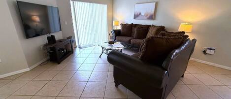 A living room with a large flat-screen TV, a dark leather sofa, a coffee table, and a dining table. There is a ceiling fan, a window with blinds, and a painting on the wall.