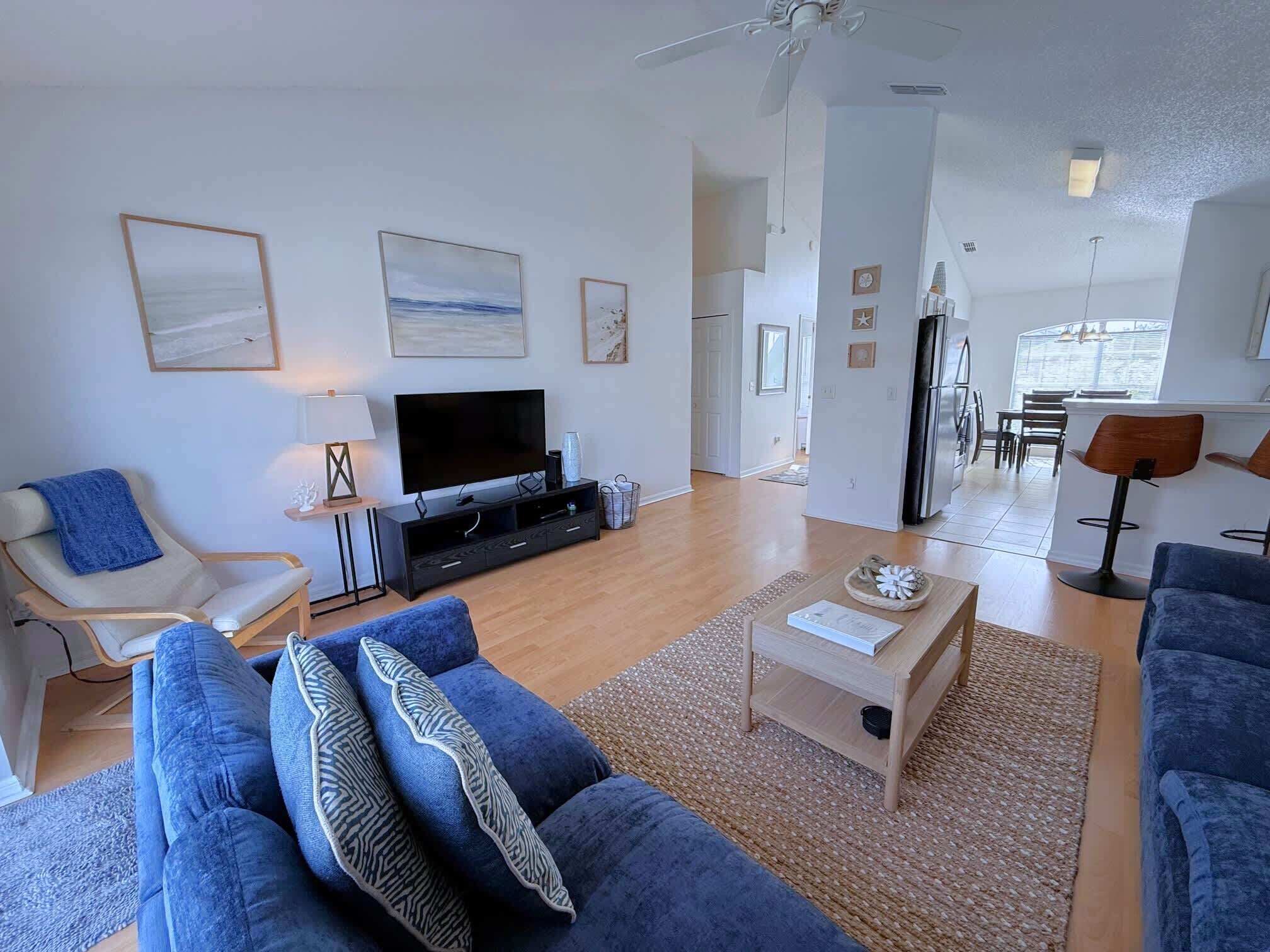 A modern living room with a flat-screen TV, blue sofas, a coffee table, and a dining area with chairs and a table.