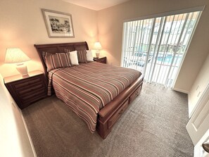 A bedroom with a large bed, two bedside tables with lamps, a framed picture on the wall, and a view of a swimming pool through the window. - Stunning Villa With Golf Course Views (Haines City)