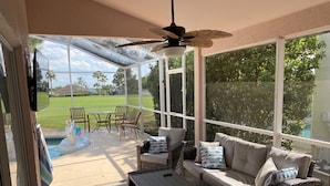 En overdækket terrasse med fletmøbler, ventilator og udsigt til swimmingpool og græsplæne.