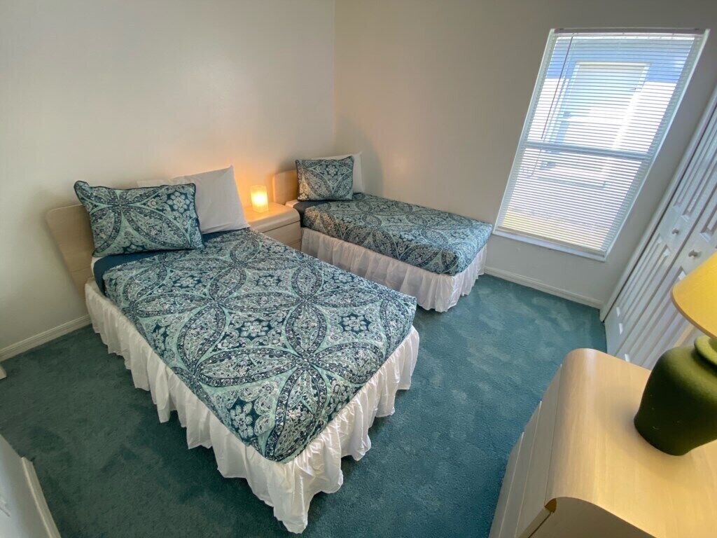 A bedroom with two beds, each with a patterned bedspread and matching pillows, a bedside table, and a window with blinds.