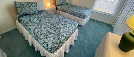 A bedroom with two beds, each with a patterned bedspread and matching pillows, a bedside table, and a window with blinds.