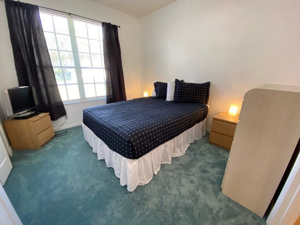 A bedroom with a bed, a television on a side table, and a window with curtains.