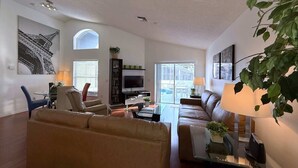 A spacious living room with a large sofa, a coffee table, and a television. There is a bookshelf, a dining area with chairs, and a large window providing natural light. - South Facing Villa - Modern Finishes (Haines City)