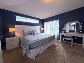 A bedroom with a bed, bedside lamps, a desk with a mirror, and a window with blinds. - South Facing Villa - Modern Finishes (Haines City)