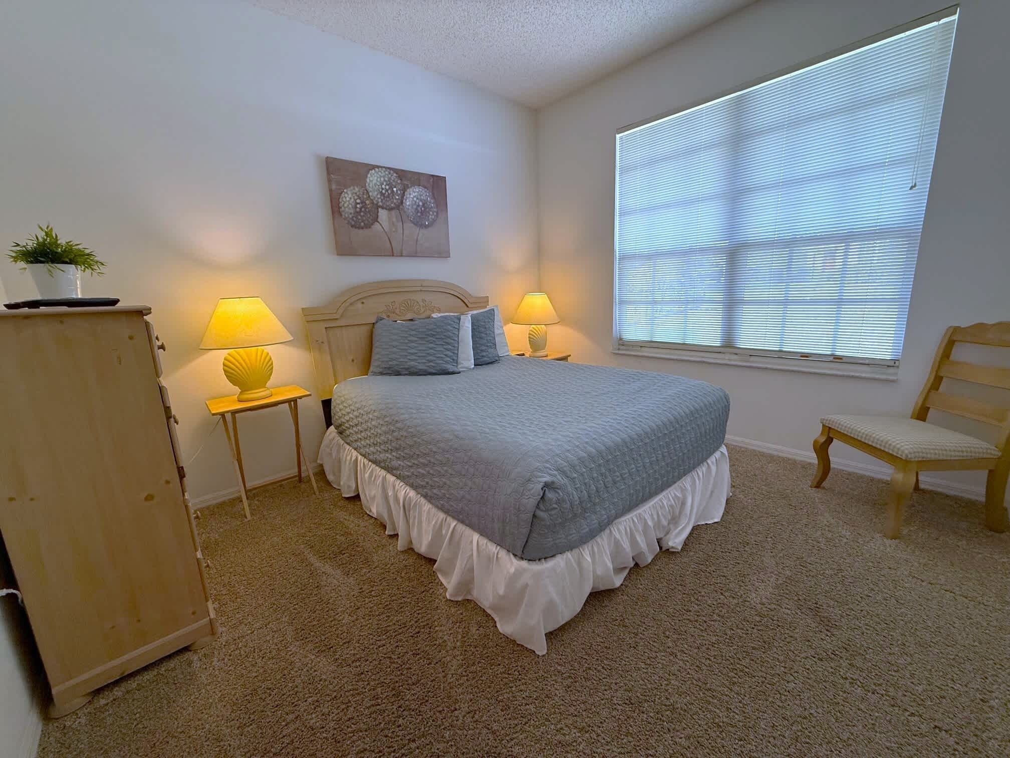 A bedroom with a bed, two bedside tables with lamps, a wooden chair, a window with blinds, and a wall-mounted artwork.