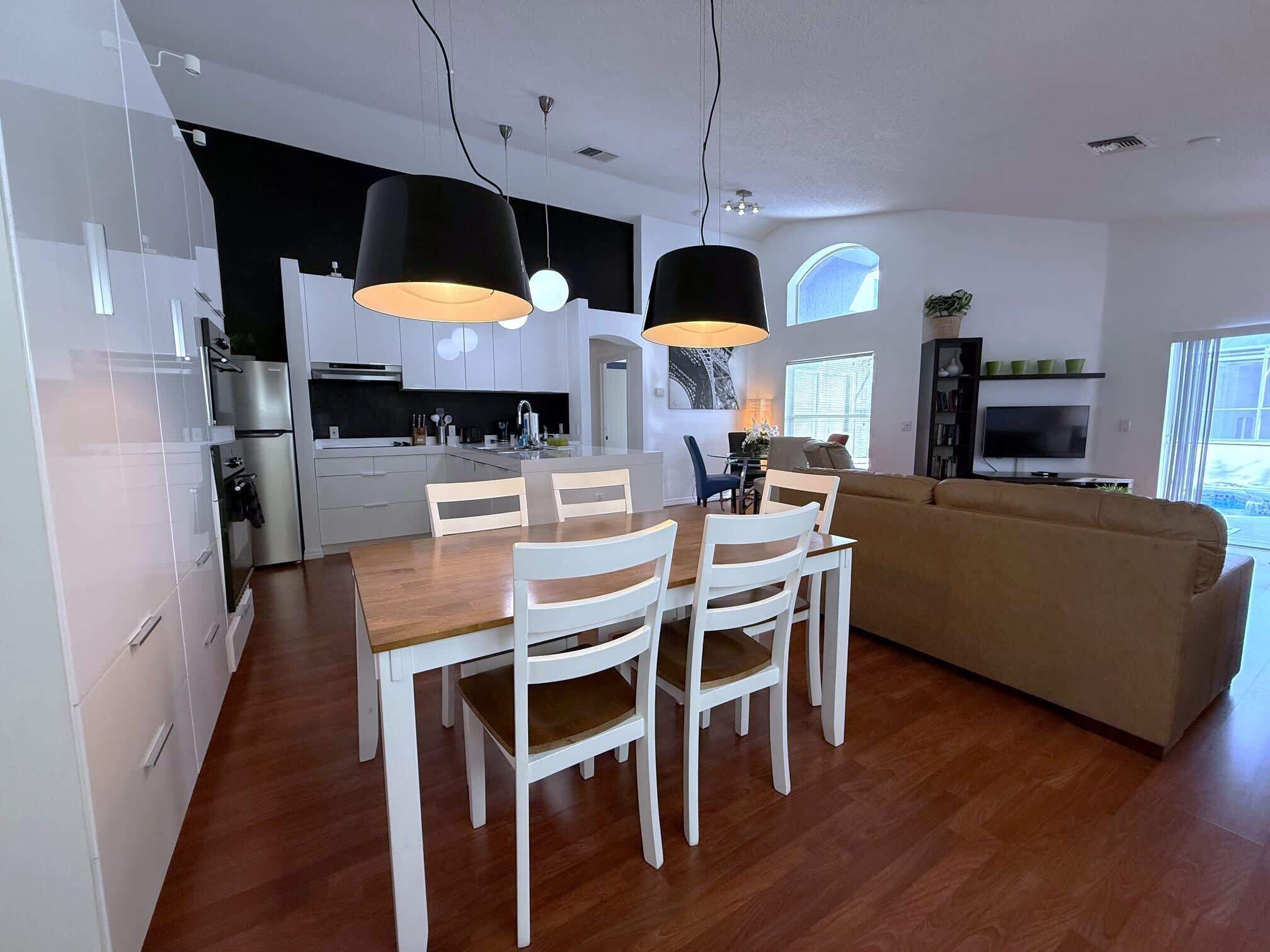 A modern kitchen with white cabinets, a dining table with chairs, a sofa, and pendant lighting.