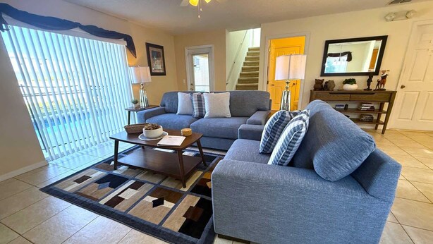 Ein Wohnzimmer mit grauem Sofa, Couchtisch und einem gemusterten Teppich. Eine Treppe führt zu einer weiteren Ebene, eine Tür und ein Fenster mit Jalousien sind ebenfalls vorhanden.