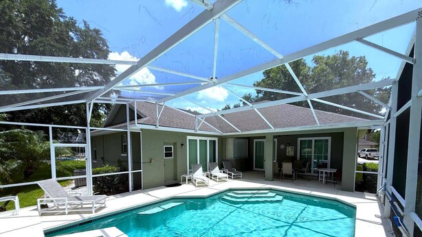 A covered swimming pool with a clear blue water surface, surrounded by a white deck and a covered patio area with chairs and a table. - Golf Course Views, Large Pool Deck Villa in Southern Dunes (Haines City)