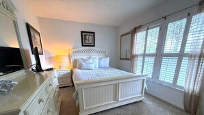Un dormitorio con una cama grande, un aparador, una mesita de noche, una lámpara, un cuadro enmarcado en la pared y una ventana con cortinas.