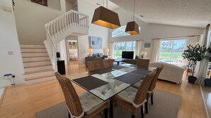 A spacious living area with a glass-top dining table, wicker chairs, a staircase, a sofa, and a large window offering a view. - 8th Fairway Pool Villa, Southern Dunes (Haines City)